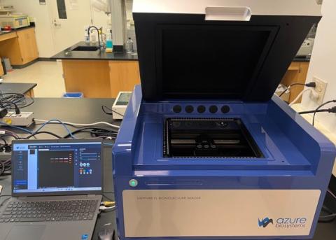 Open laboratory instrument with lid raised next to a laptop on a lab bench.